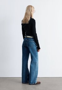 Schwarzes, geripptes Langarmoberteil, kombiniert mit hoch taillierten, weiten blauen Jeans aus Denim; ein gemusterter Gürtel ist an der Taille sichtbar.