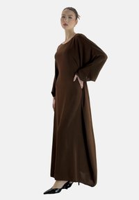 Robe longue, marron, fluide avec des manches amples et un col rond, assortie à des talons noirs pointus. Tissu lisse avec une texture douce.