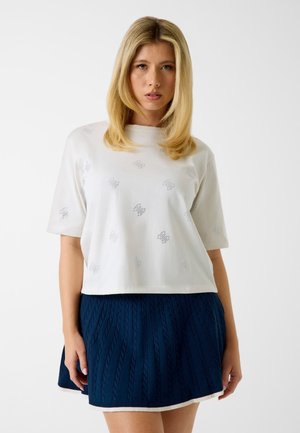 Femme blonde portant une chemise blanche à manches courtes avec de petites impressions papillons et une mini-jupe texturée bleu marine, debout sur un fond blanc.