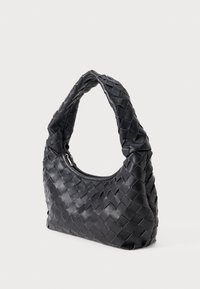 Musta kudottu nahkalaukku olkahihnalla, jossa on pehmeä ja paksu hihna sekä ylhäältä avattava vetoketju valkoista taustaa vasten.