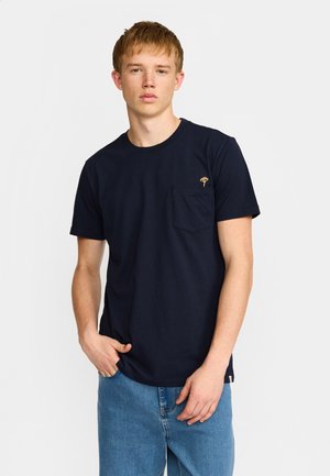 Jonge man die een navyblauw T-shirt met korte mouwen draagt met een klein geborduurd embleem op het borstzakje en lichtblauwe spijkerbroek.