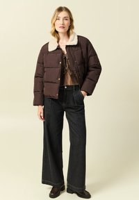 Giacca a vento marrone con colletto foderato in pelliccia, abbinata a un top marrone e jeans scuri a gamba larga. La modella indossa stivaletti neri.