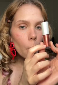 Lipgloss in een doorzichtig tubetje met een zilveren dop, in een metallic rosegoud kleur. Achtergrond vervaagd, focus op het product dat in de handen wordt gehouden.