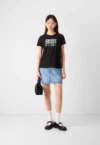 T-shirt nera con logo "DKNY" argento, abbinata a una gonna di jeans blu chiaro. Gli accessori includono una borsa nera e scarpe nere con cinturini.