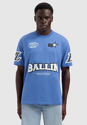 Man draagt een blauwe "BALLIN" grafische t-shirt en lichte denim jeans, kijkt recht vooruit met een neutrale uitdrukking tegen een effen achtergrond.