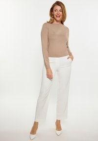 Beige langärmliges Oberteil mit Rippstruktur und Rundhalsausschnitt, kombiniert mit weißen maßgeschneiderten Hosen. Das Model posiert mit den Händen in den Taschen und trägt weiße High Heels.