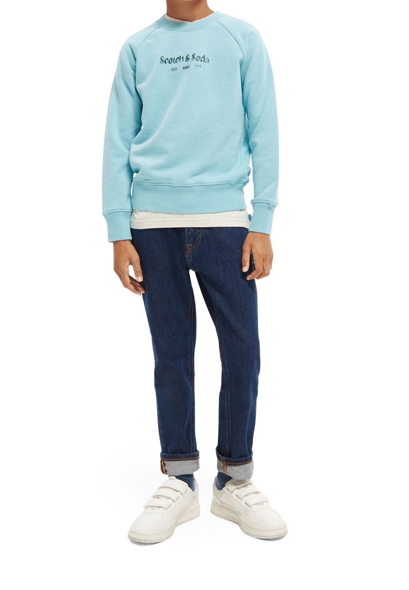 Sudadera azul claro con mangas raglán, logo en el pecho, superpuesta a una camisa blanca. Combinada con jeans oscuros y zapatillas blancas.