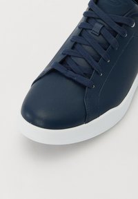 Baskets en cuir bleu marine avec une texture lisse, semelle en caoutchouc blanc, lacets plats et design minimaliste. Les accents sont simples et épurés.
