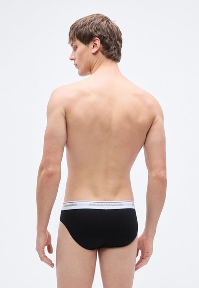 Slips en coton noir avec une texture côtelée et une ceinture blanche à logo, vus de dos, mettant en avant la coupe et le design.