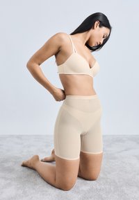 Pantaloni scurți bej modele fără cusături, cu talie înaltă, proiectați pentru o potrivire perfectă. Model ajustând potrivirea în timp ce îngenunchează pe o suprafață texturată.