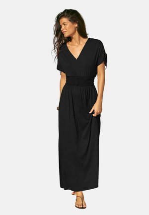 Naine kannab musta V-kaelusega maksikleit, millel on volangid vöökohal ja lühikesed varrukad, seisab ning vaatab küljele, hoides ühe käega kleiti.