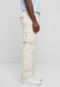 Pantalons cargo beiges à coupe loose avec plusieurs poches latérales, coutures visibles et ourlet ajustable. Associés à des baskets blanches.