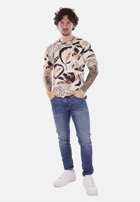 Camisa de manga corta con base crema y llamativos patrones de hojas en marrón y negro, combinada con jeans azules y zapatillas blancas.