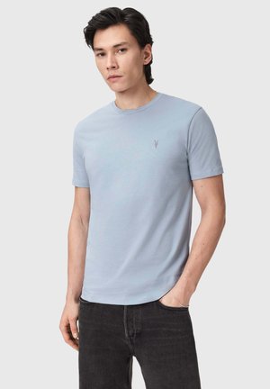Ung mand iført en lyseblå t-shirts med korte ærmer og et lille broderet logo på brystet samt sorte jeans, poserer med den ene hånd i lommen.
