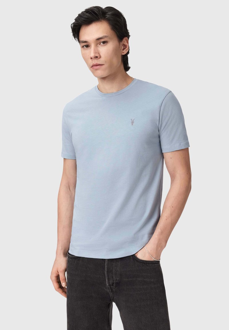 Jeune homme portant un t-shirt à manches courtes bleu clair avec un petit logo brodé sur la poitrine et un jean noir, posant avec une main dans la poche.