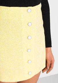 Jupe en tissu jaune avec un tissage texturé, ornée de boutons ronds blancs bordés d'or, et une longueur juste au-dessus du genou.