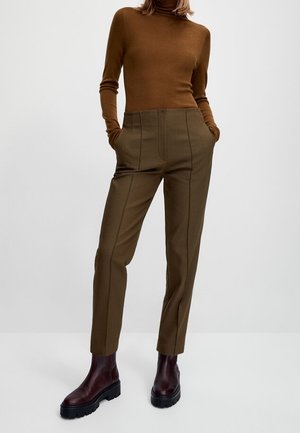 Personne portant un col roulé marron ajusté, un pantalon sur mesure taille haute vert olive et des bottines marron foncé, debout les mains dans les poches.
