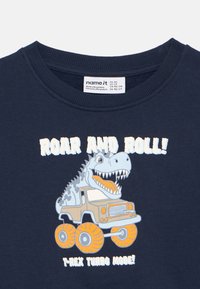 Ναυτικό μπλε μπλουζάκι με γραφικό ενός Τ-ρεξ σε φορτηγάκι με πορτοκαλί τροχούς, κείμενο "ROAR AND ROLL!" σε λευκό και απαλό μπλε. Υφασμάτινο από βαμβάκι.