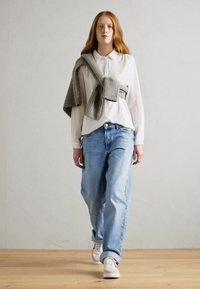 Weißes Hemd mit Knöpfen, hellblaue lockere Jeans, grauer Pullover über den Schultern, weiße Sneakers, Holzboden, schlichter heller Hintergrund.