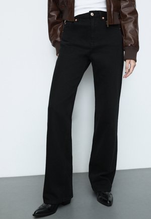 Persoon draagt zwarte, hoge taille, wijde broekspijpen, een bruine leren jas, een wit topje en zwarte puntige schoenen, staand tegen een lichte muur.