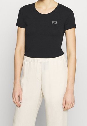 Person iført en sort tætsiddende T-shirt med "Liu Jo Sport"-logo på brystet og løstsiddende cremefarvede højtaljede bukser.