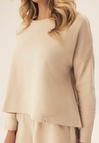 Kremowy, luźny sweter z szerokim dekoltem i długimi rękawami, z małą metką z logo na dolnym brzegu. Miękka tekstura.