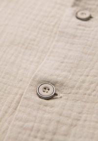Gros plan d’un tissu beige texturé avec deux boutons blancs cousus, un net près du centre et un flou à l’arrière-plan.