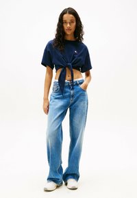 Marineblauw cropped t-shirt met een knoop aan de voorkant, gecombineerd met losse blauwe spijkerbroeken. Opvallend logo-detail op het shirt, casual ontwerp, katoenen stof.