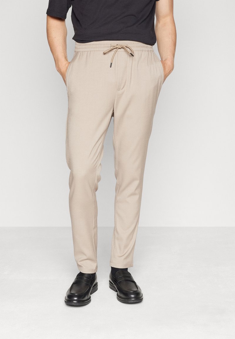 Beiga, avsmalnande byxor med elastiskt midjeband och dragsko. Har sidfickor och en smidig textur. Matchas med svarta loafers.