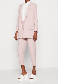 Lichtroze op maat gemaakte blazer en bijpassende cropped broek, gecombineerd met chunky witte slide-sandalen. De stof lijkt glad met een gestructureerde pasvorm.