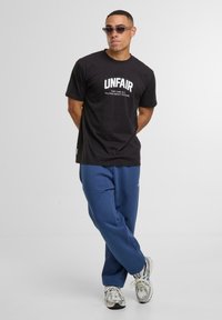 Zwarte katoenen T-shirt met witte tekst "UNFAIR" en kleinere tekst eronder. Gecombineerd met blauwe sweatpants en grijze sneakers.
