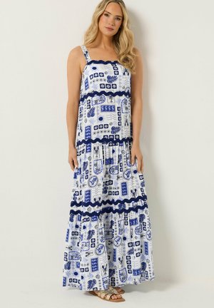 Frau, die ein ärmelloses, langes weißes Kleid mit blauen abstrakten Mustern und gezackten dunkelblauen horizontalen Streifen trägt, steht vor einer blassen Wand.