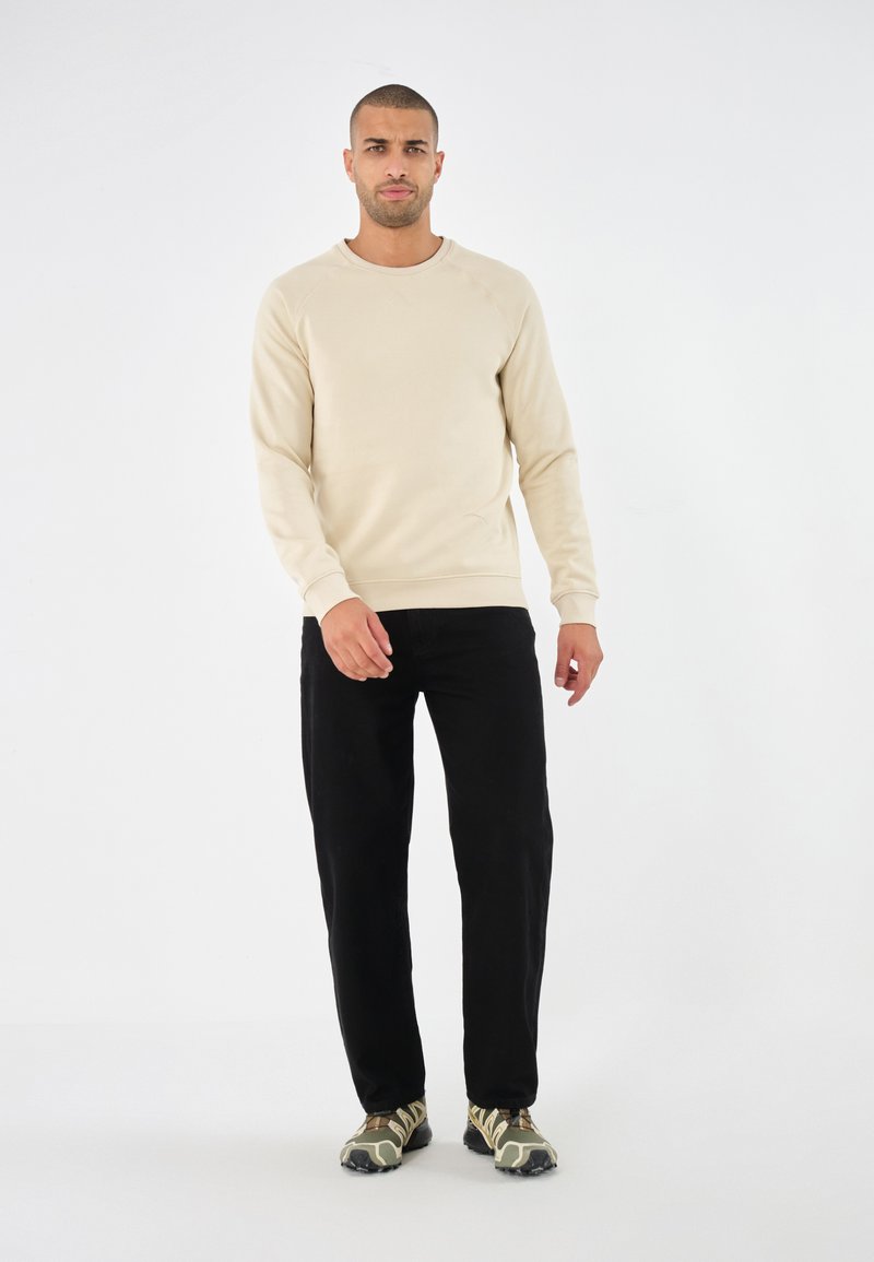 Beige sweatshirt met ronde hals, gecombineerd met zwarte broek en gestreepte schoenen. De stof lijkt zacht en iets getailleerd.