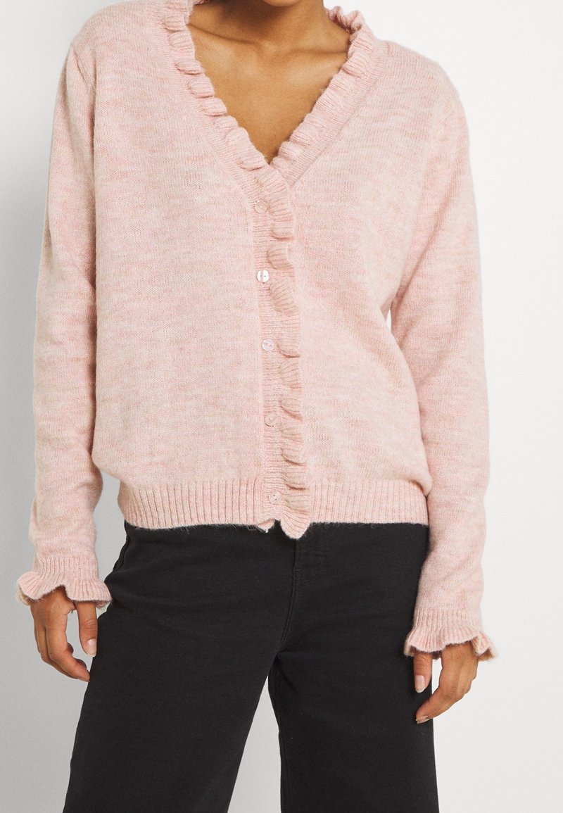 Osoba v světle růžovém cardiganovém svetru s volány, zapnutém vpředu, se černými kalhotami, s odkrytou střední částí těla a uvolněnými rukama po stranách.