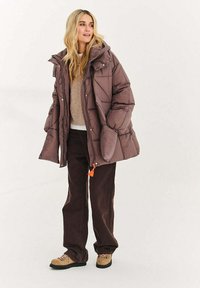 Brązowa ocieplana kurtka puchowa z kapturem, teksturowanym materiałem, zatrzaskami i elastycznymi troczkami, w połączeniu z ciemnymi spodniami i beżowymi butami.