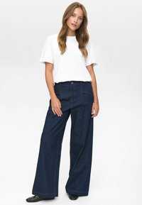 Woman with long hair wearing a plain white t-shirt, dark wide-leg jeans, and black woven slip-on shoes, standing against a white background.