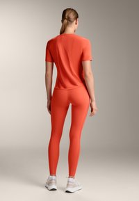 Oranžna športna majica s kratkimi rokavi in zaobljenim robom, v kombinaciji z usklajenimi leggingi, ki se odlikujejo po elegantnih linijah in podrobnostih šivanja.