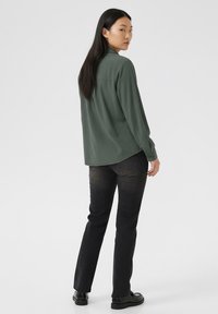 Tmavozelená košeľa s dlhým rukávom a golierom na gombíky, skombinovaná s čiernymi džínsami a čiernymi topánkami. Tkanina vyzerá hladko a má uvoľnený strih.