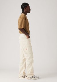 Camiseta beige, pantalones cargo blancos de corte suelto con bolsillos y zapatillas grises con detalles negros. Modelo posando de perfil contra un fondo neutro.