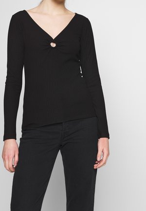 Kobieta ubrana w czarną prążkowaną bluzkę z długim rękawem, z dekoltem w kształcie litery V i okrągłym wycięciem na wysokości klatki piersiowej, w połączeniu z czarnymi spodniami.