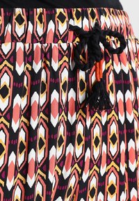 Jupe à motifs présentant des formes géométriques en orange, blanc et noir. Taille à cordon avec détail pompon, fabriquée en tissu léger.