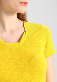 Žluté tričko s krátkým rukávem a kulatým výstřihem, vyrobené z měkké, texturované tkaniny, s mírně volným střihem a viditelnými švy.