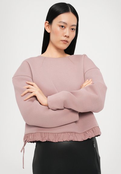 Roosa lühike dressipluus rippuva äärisega, siledast materjalist ja pikkade õhukeste varrukatega. Kombineeritud musta seeliku ning tõmbluku detailiga.