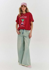 Czerwona, oversize'owa koszulka z nadrukiem graficznym, z podwiniętymi rękawami, w połączeniu z jasnoniebieskimi spodniami typu wide-leg z postrzępionymi końcami oraz różową czapką.