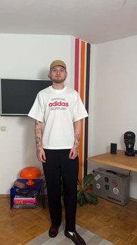 Balta īsroku T-krekls ar sarkanu un melnu uzrakstu "oficiālais adidas piegādātājs", kombinēts ar melnām biksēm un tumšiem apaviem.