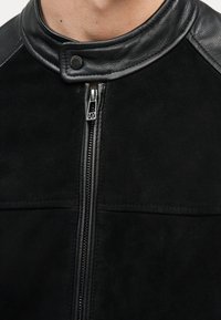 Κοντινό πλάνο μαύρου μπουφάν με δερμάτινο γιακά και πάνελς στους ώμους, με κεντρικό ασημένιο φερμουάρ που έχει το λογότυπο AX στην ταινία έλξης και κλείσιμο με κουμπί ασφαλείας.