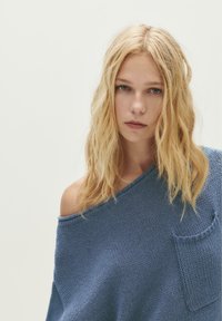 Blonde Frau mit welligem Haar, die einen lockeren blauen Strickpullover trägt, der von einer Schulter rutscht, und direkt in die Kamera schaut, vor einem schlichten hellen Hintergrund.