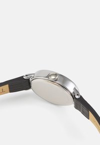 Boîtier de montre en acier inoxydable argenté avec un dos lisse, mettant en avant un bracelet noir texturé et une couronne métallique proéminente sur le côté.