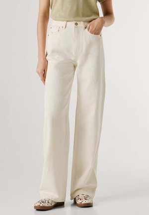 Personne portant un pantalon blanc taille haute à jambes larges, une main dans la poche, associé à des chaussures slip-on tachetées et une chemise vert clair.