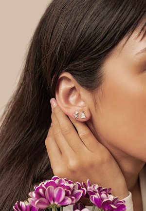 Silver stud earrings featuring a gingerbread man and Christmas tree design. Close-up view with a hand near the ear and flowers in the foreground.